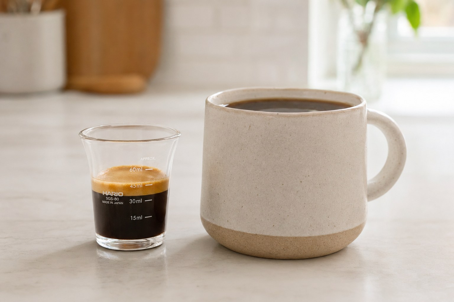 Hario SGS-80 espresso shot glass next to a full ceramic mug of drip coffee on a kitchen counter, side by side for size comparison